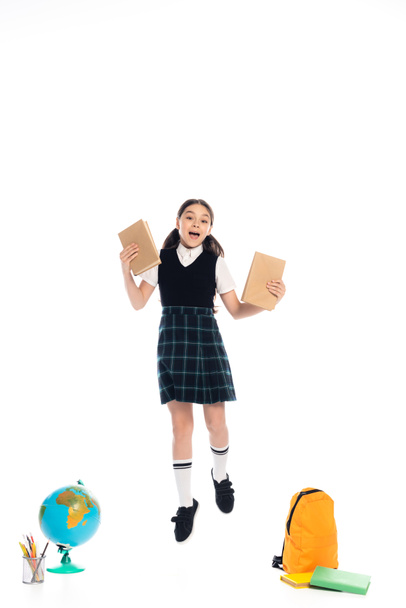 Excited schoolkid holding books while jumping near globe and backpack on white background - Photo, Image