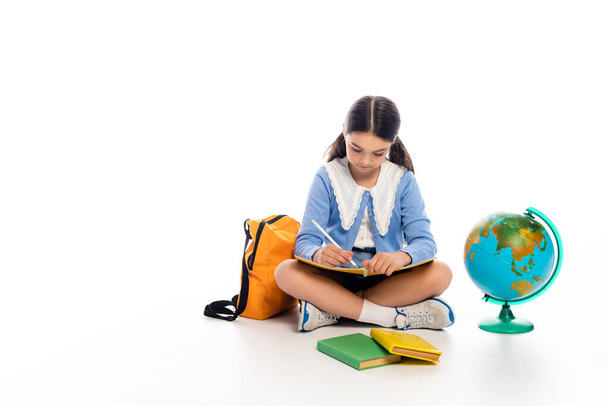 Preteen schoolkid writing on notebook near backpack and globe on white background - Photo, Image