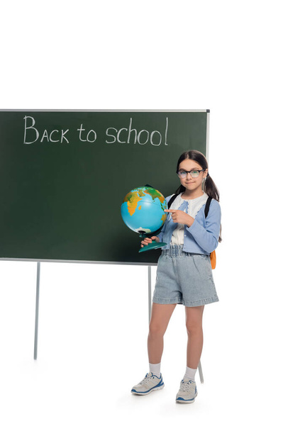 Usmívající se školák ukazuje na zeměkouli v blízkosti tabuli s zpět do školy nápisy na bílém pozadí - Fotografie, Obrázek