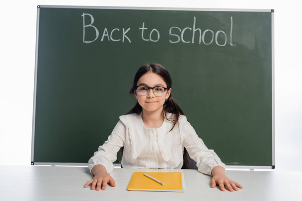 Улыбающаяся школьница смотрит в камеру возле блокнота и доски со школьными надписями, изолированными на белом  - Фото, изображение