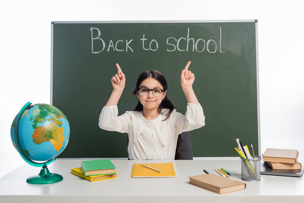 Glimlachend schoolkind wijzend naar schoolbord met terug naar school belettering bijna wereld en boeken geïsoleerd op wit  - Foto, afbeelding