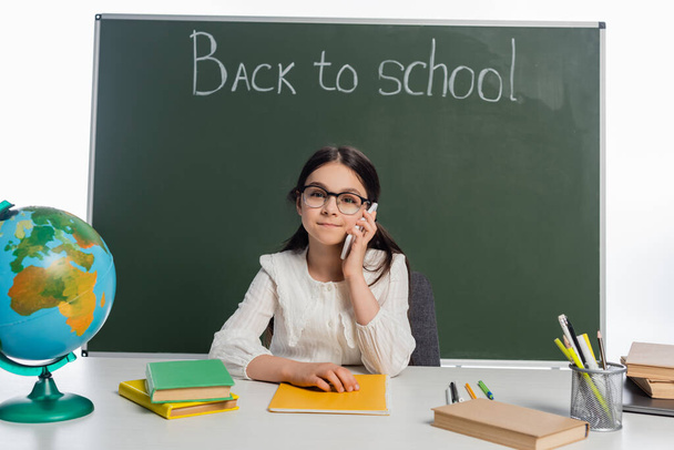 Pupil talking on smartphone near books and chalkboard with back to school lettering isolated on white  - Photo, Image