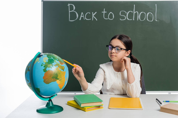 Schoolgirl pointing at globe near books and chalkboard with back to school lettering isolated on white  - 写真・画像