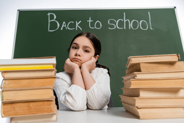 Schoolkid looking at camera near blurred books and chalkboard with back to school lettering isolated on white  - Foto, imagen