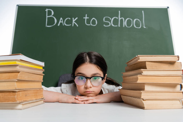Overstuur schoolmeisje in bril kijken naar camera in de buurt van boeken en schoolbord met terug naar school belettering geïsoleerd op wit  - Foto, afbeelding