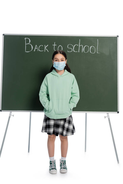 Tıp maskeli okul çocuğu beyaz arka planda yazı tahtasının yanında duruyor. - Fotoğraf, Görsel