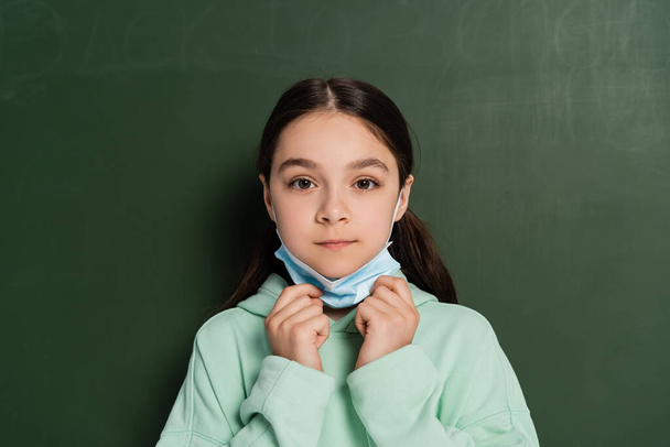 Schoolgirl holding medical mask near chalkboard at background - Foto, Imagem