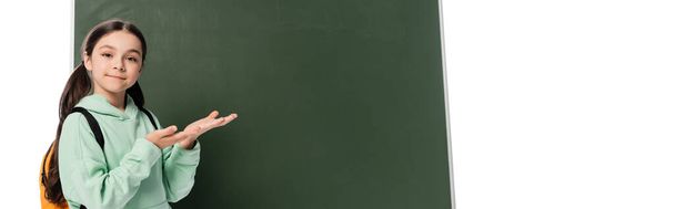Escolar sonriente con mochila apuntando a pizarra aislada en blanco, pancarta  - Foto, imagen