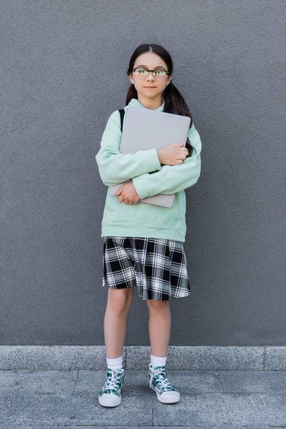 Pupil in eyeglasses holding laptop near building outdoors  - Photo, Image