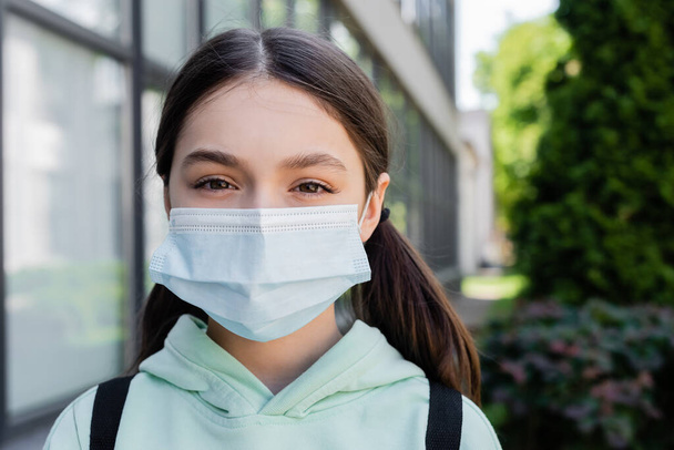 Portrait of schoolgirl in medical mask looking at camera on urban street  - Photo, Image