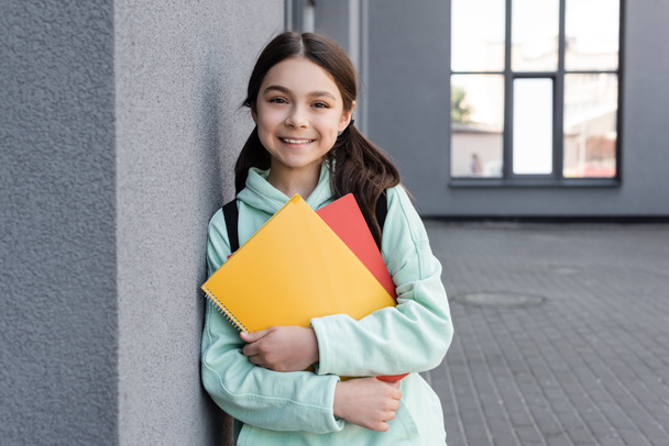 Cheerful schoolgirl holding notebooks and looking at camera near building outdoors  - Photo, Image