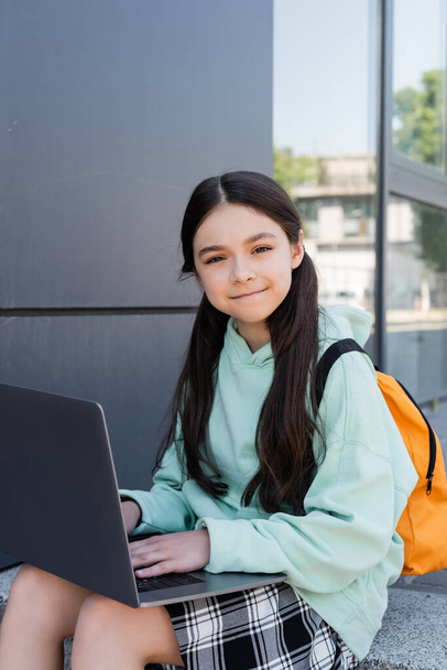 Positive schoolgirl with backpack using laptop near building outdoors  - 写真・画像