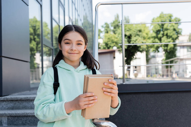 Escolar sonriente con mochila sosteniendo libros cerca del edificio al aire libre  - Foto, Imagen