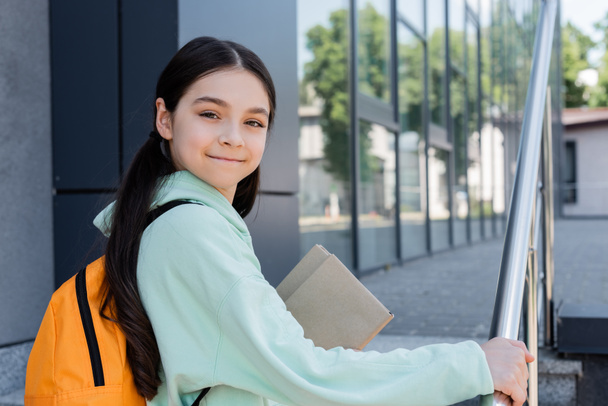 Positive pupil with backpack and books looking at camera near blurred books outdoors  - Photo, Image