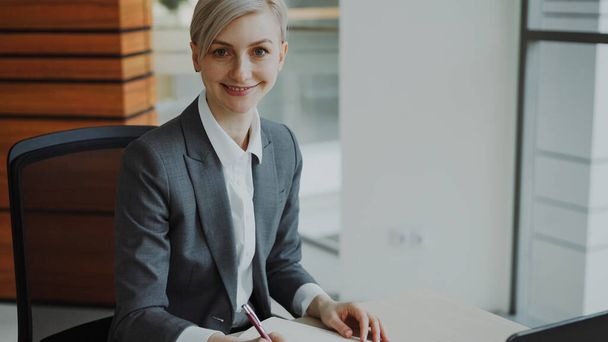 Portrait of Attractive blonde businesswoman sitting at table writing in notebook smiling into camera in modern office indoors - Foto, afbeelding