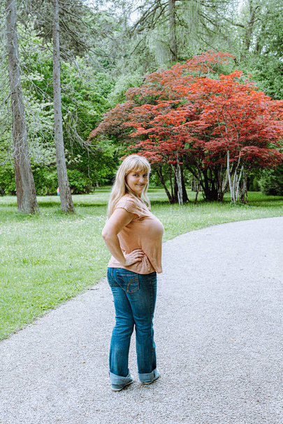 Atraktivní zakřivená mladá žena, v neformálních džínách a tričku, blond s dlouhými vlasy, dlouhá na pěšině parku. portrét ženy na pozadí krajinářského parku, při pohledu do kamery - Fotografie, Obrázek