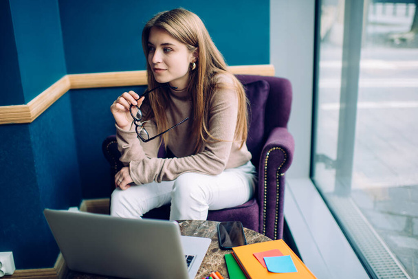 Positive young female in casual wear sitting in comfortable armchair near table with laptop and smartphone while holding glasses and looking away - Fotoğraf, Görsel