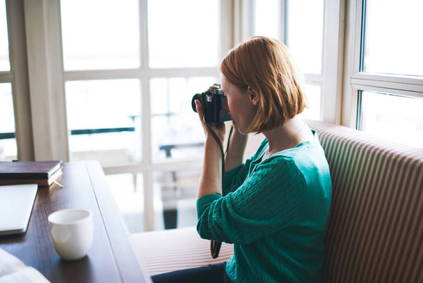 Side view of focused female photographer in casual clothes sitting at table with cup of coffee and taking photo in cafeteria - Фото, изображение