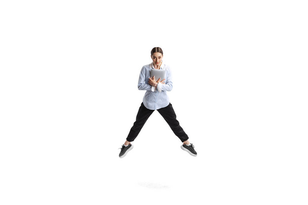 Portret van een jonge vrouw, kantoormedewerker in officiële outfit springend met laptop geïsoleerd over witte studio achtergrond. Concept van het bedrijfsleven, kantoor levensstijl, succes, ballet, carrière, expressie, advertentie - Foto, afbeelding