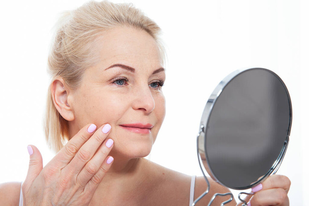 Happy middle aged woman model touching face skin looking in mirror. Smiling mature older lady enjoying healthy skin care. Aging beauty, skincare treatment cosmetic products concept. - Fotoğraf, Görsel