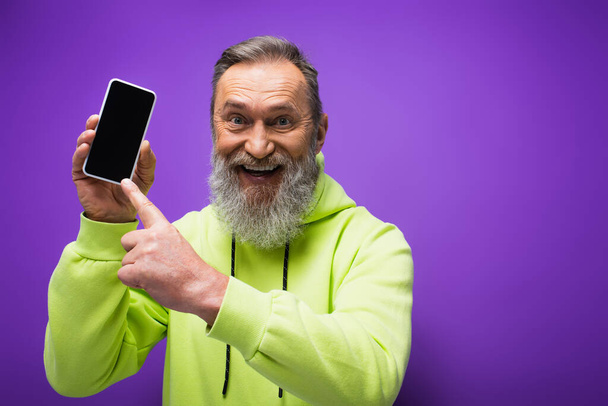 happy senior man with beard and grey hair pointing at smartphone on purple  - Fotó, kép