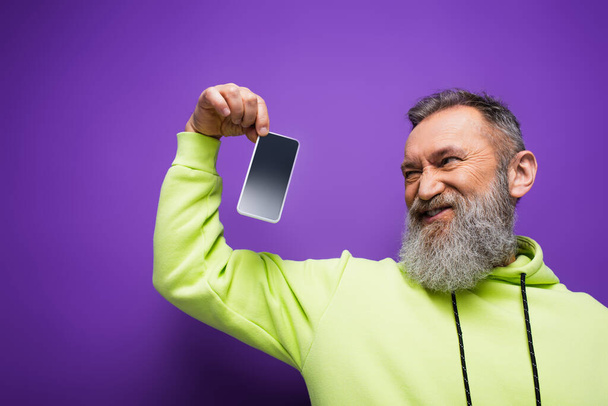displeased senior man with beard and grey hair holding smartphone with blank screen on purple  - Foto, Bild