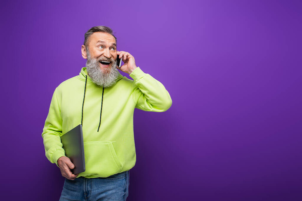 happy senior man with beard and grey hair holding laptop while talking on mobile phone on purple  - Фото, зображення