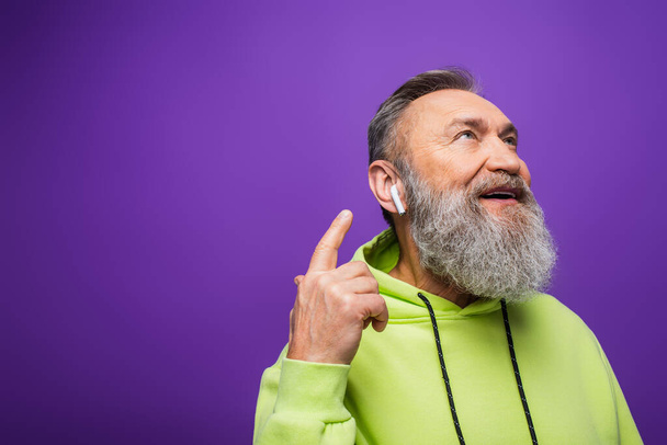 Yeşil kapüşonlu mutlu adam müzik dinliyor ve mor kulaklıklı kablosuz kulaklığı işaret ediyor.  - Fotoğraf, Görsel