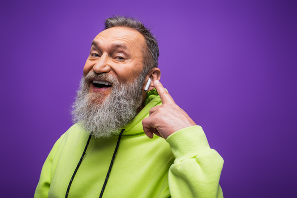 cheerful senior man in green hoodie listening music and pointing with finger at wireless earphone on purple - Fotografie, Obrázek