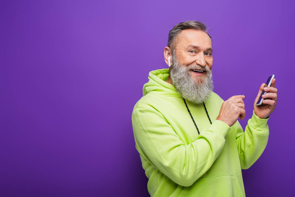 happy senior man in green hoodie listening music in wireless earphone and holding smartphone on purple  - Fotó, kép