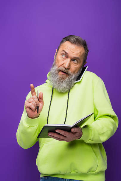 senior man in green hoodie holding pen and notebook while talking on smartphone on purple  - Foto, imagen