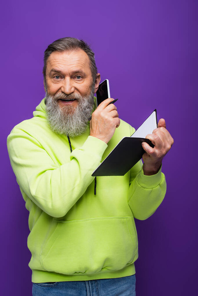 glücklicher Senior in grünem Kapuzenpulli hält Notizbuch in der Hand und telefoniert auf dem Smartphone lila  - Foto, Bild
