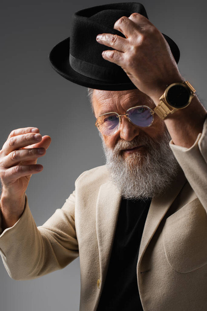 elegant senior man in beige jacket and eyeglasses wearing derby hat on grey - 写真・画像
