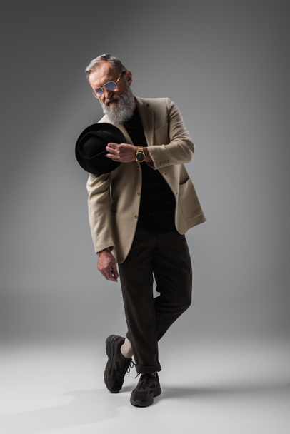 full length of elegant senior man in beige blazer and eyeglasses posing with derby hat on grey - 写真・画像
