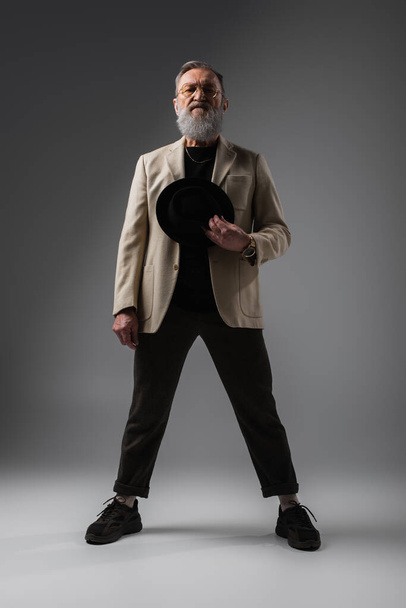 full length of elegant senior man in beige jacket and eyeglasses standing with derby hat on grey - Valokuva, kuva