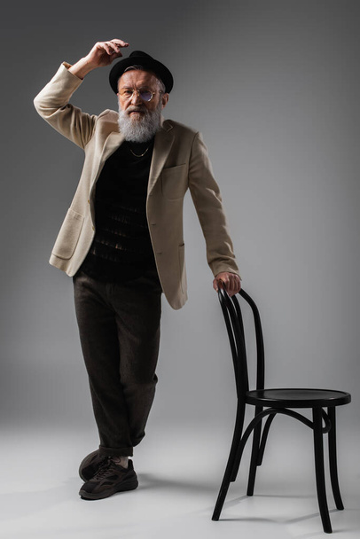 full length of elegant senior man in beige jacket and eyeglasses posing with derby hat near chair on grey - Photo, Image