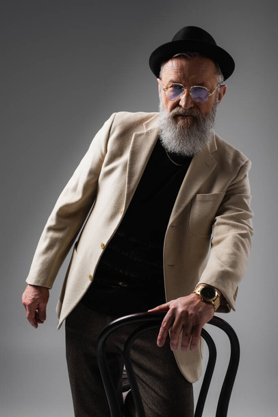 elegant senior man in beige jacket and eyeglasses posing with derby hat near chair on grey - Φωτογραφία, εικόνα