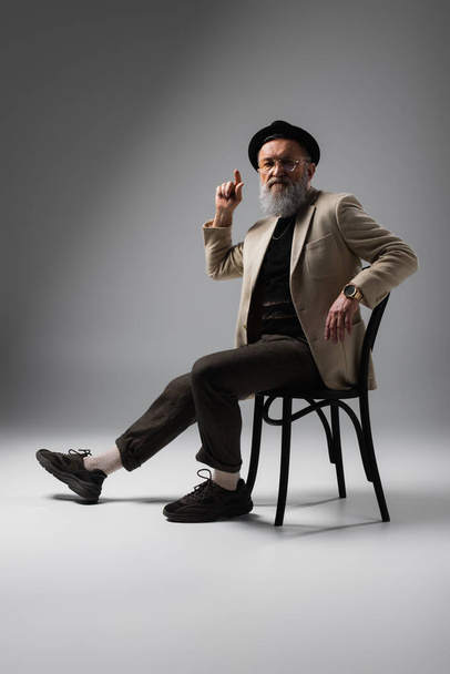 full length of elegant senior man in beige jacket and derby hat sitting on wooden chair on grey - Fotoğraf, Görsel