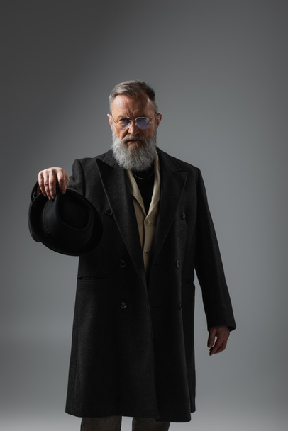 stylish senior man in dark coat and eyeglasses holding derby hat on grey - Fotografie, Obrázek