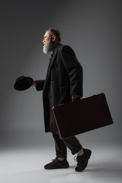 full length of elegant senior man in stylish coat holding derby hat and suitcase on grey - Foto, Imagem