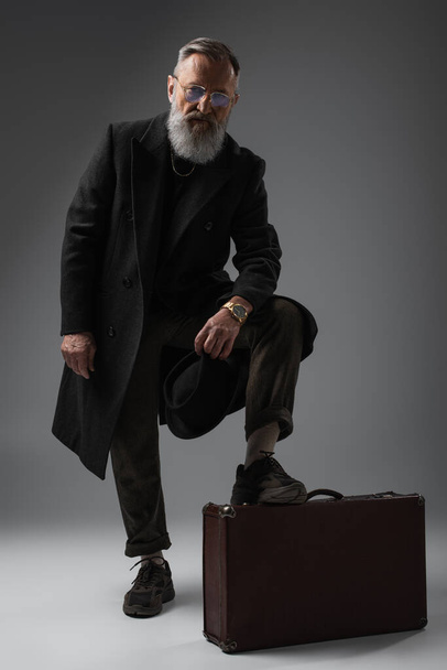 full length of elegant senior man in stylish coat holding derby hat and putting leg on  suitcase on grey - 写真・画像
