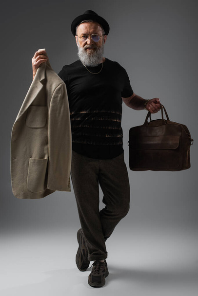 full length of elegant senior man in derby hat holding leather bag and beige jacket on grey - Valokuva, kuva