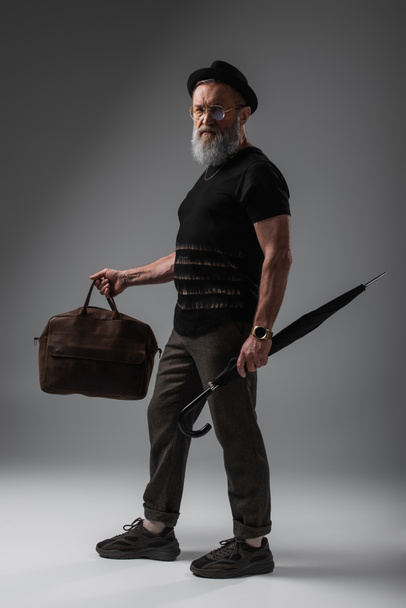 full length of bearded senior man in derby hat holding leather bag and umbrella on grey - 写真・画像