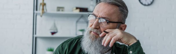 töprengő és szakállas idős férfi szemüvegben tollal a kezében, és néz el otthonról, banner - Fotó, kép