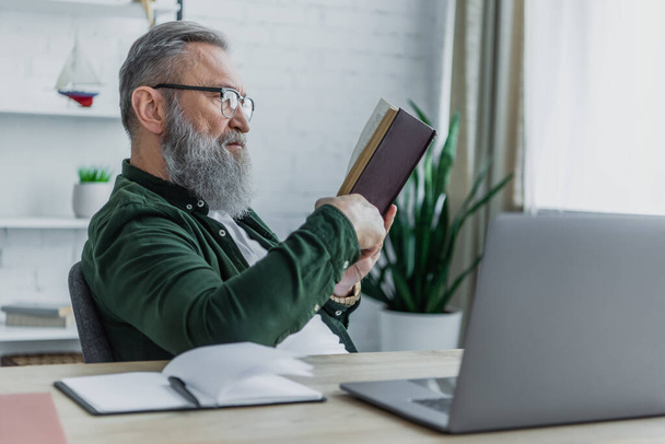 bearded senior man in eyeglasses reading book near laptop on desk - Zdjęcie, obraz