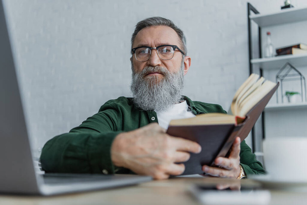 bearded senior man in eyeglasses looking at camera near laptop while holding book - Фото, изображение