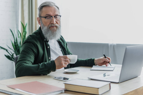 szakállas idősebb férfi szemüvegben kezében csésze kávé és toll mellett laptop, miközben dolgozik otthonról  - Fotó, kép