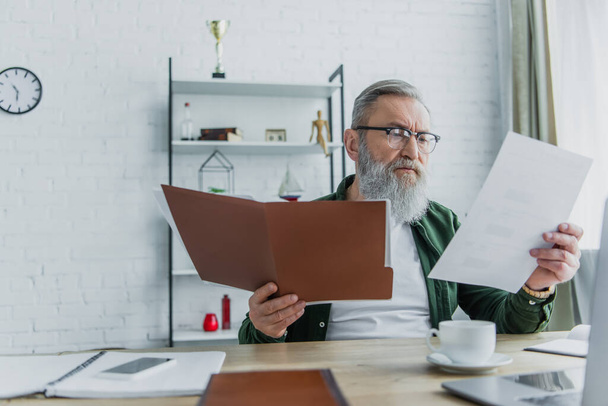 старший человек в очках держит папку и смотрит на документ рядом с ноутбуком на столе - Фото, изображение