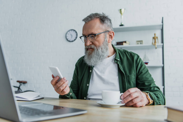 bearded senior man in eyeglasses holding cup and looking at smartphone near laptop on desk  - Fotó, kép