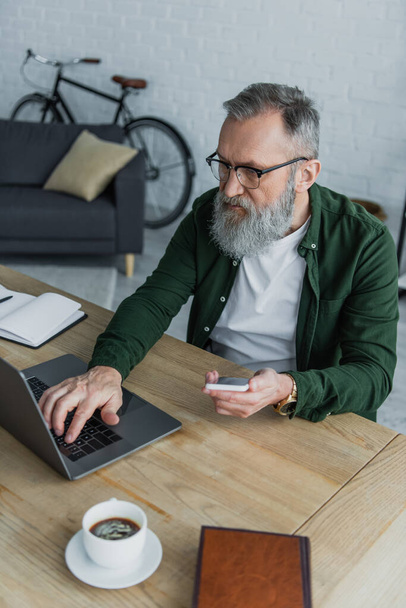 bearded senior man in eyeglasses holding smartphone while using laptop at home - Fotoğraf, Görsel